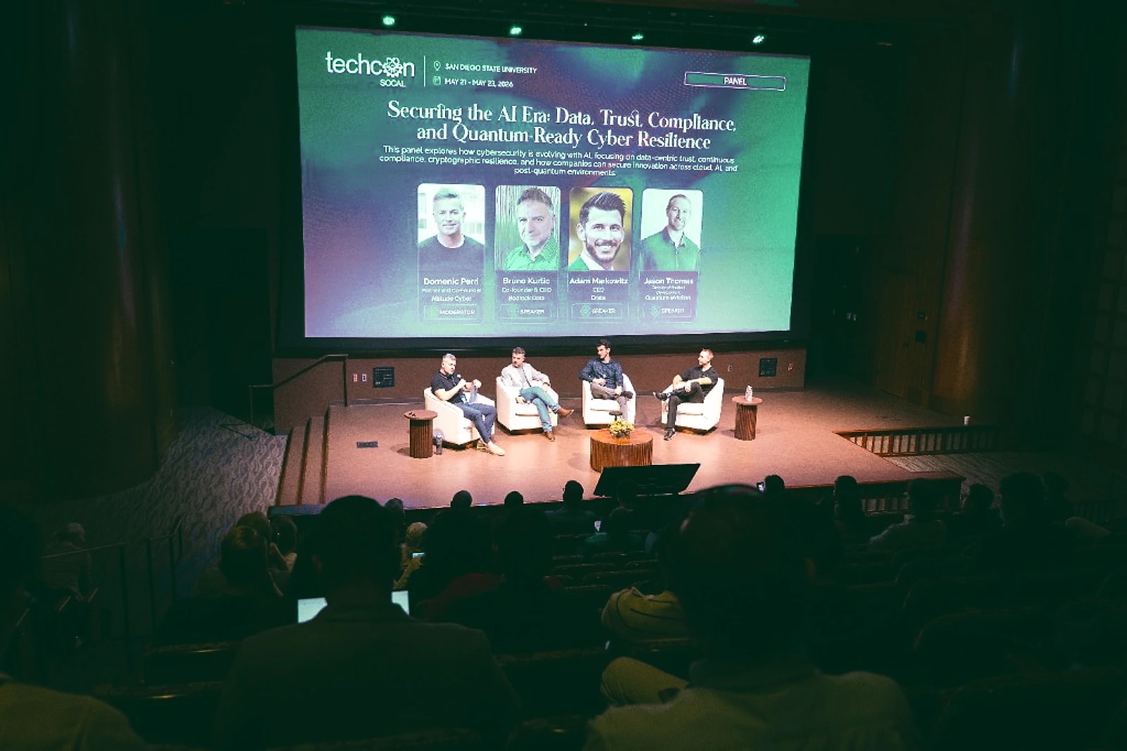 Four panelists in conversation on stage beneath a large presentation screen, facing a seated audience